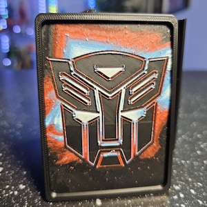 May include: A rectangular black frame containing a raised, black Autobot insignia from the Transformers franchise. The background behind the logo features a textured, swirling blend of vibrant red and bright blue colors, creating a high-contrast effect.