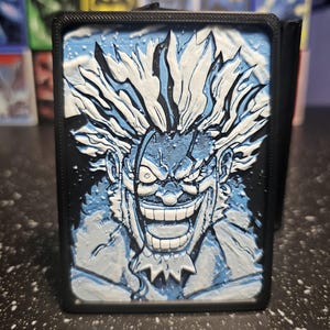 May include: A rectangular black plastic deck box featuring a 3D-printed relief of a spiky-haired character with a wide, toothy grin. The character is rendered in shades of blue and white, set against a dark background within a black frame.