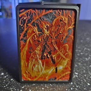 May include: A black card case featuring a fiery, embossed design of a phoenix. The artwork is predominantly orange and yellow against a black background, with intricate details. The case has a rectangular shape.
