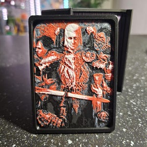 May include: A black rectangular case with a detailed relief image in red and black. The image depicts several figures, including a central character with white hair, holding a sword. The case is likely for a card or small item.