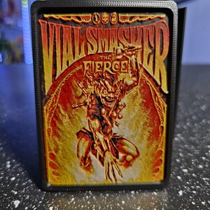 May include: A rectangular black frame containing a 3D-printed card featuring a vibrant illustration of a goblin-like character. The text VIAL SMASHER THE FIERCE is printed in bold yellow and orange letters above the central figure in a fiery setting.