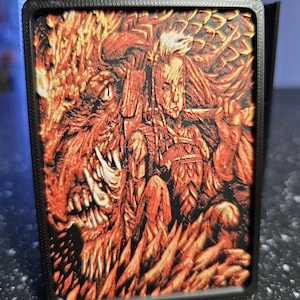 May include: A rectangular black frame containing a 3D-printed relief panel with a textured surface. The artwork features a detailed scene in shades of orange, red, and black, depicting a monstrous creature with sharp teeth alongside a human figure.