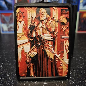 May include: A rectangular 3D-printed card case featuring a relief illustration of a bearded knight in ornate armor holding a sword. The knight stands within a red-toned interior setting. The case has a solid black frame and a textured surface.