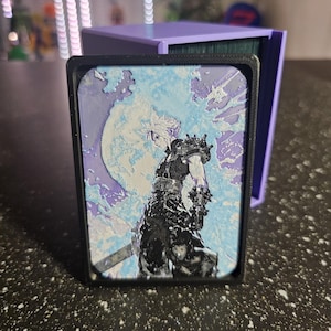May include: A black card holder with a Final Fantasy VII design. The card features a character in black armor against a blue and white background with a large moon. A purple card holder is behind it.