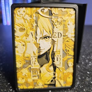 May include: A rectangular black frame containing a multi-layered 3D relief art piece in shades of yellow and cream. The artwork features a central portrait of a character surrounded by smaller vignettes of the same figure, with the text WANTED visible.