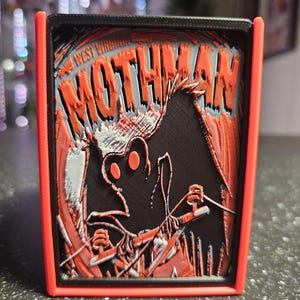 May include: A 3D-printed rectangular sign with a red frame and a black interior. The sign features the text "WEST VIRGINIA MOTHMAN" in orange and gray, along with a stylized Mothman illustration. The design is layered, creating a 3D effect.