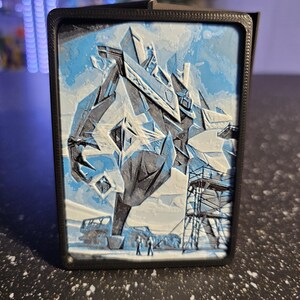May include: A rectangular black frame containing a multi-layered 3D relief print. The artwork features a large, angular, crystalline structure in shades of blue, white, and black, with small figures standing near scaffolding at the base.