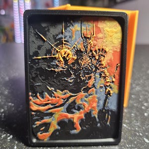 May include: A black rectangular object with a detailed, colorful relief of a fantasy figure. The artwork features a dark, textured background with orange, yellow, and blue accents. The figure is depicted with a crown and armor, set against a fiery backdrop.