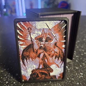 May include: A fantasy-themed card with a black border, featuring a detailed illustration of a winged figure holding a staff and sword. The artwork is predominantly red and white, set against a purple and white background. A black card storage box is in the background.