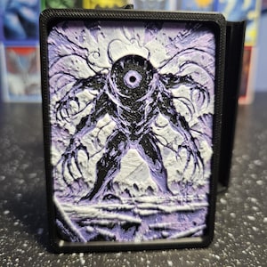 May include: A rectangular black plastic deck box featuring a relief-style lid with a central cyclopean creature design. The figure is rendered in deep black with purple and white highlights, showcasing long clawed limbs and a single central eye.