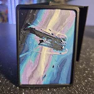 May include: A black card holder with a colorful, textured image of a spaceship in a nebula. The artwork features shades of blue, purple, and white, creating a cosmic scene. The card holder is made of a matte black material.