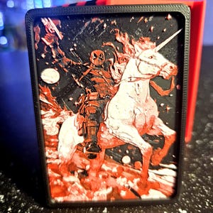 May include: A black and red rectangular case featuring a Deadpool-themed design. The artwork depicts Deadpool riding a white unicorn, set against a black background with red accents. The case has a black border and a red side.