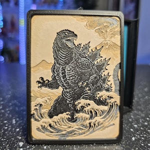 May include: A rectangular black-framed wall art piece featuring a relief-style illustration of a black Godzilla figure standing amidst stylized ocean waves. In the background, a mountain with a plume of smoke is depicted in a light tan color.