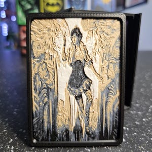 May include: A rectangular black frame containing a multi-layered relief art piece. The central image features a figure with wings, flanked by two standing figures, rendered in shades of gold, white, and dark blue with intricate carved textures.