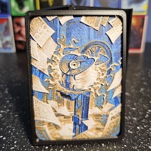 May include: A black rectangular deck box featuring a multi-layered relief art panel on the front. The artwork depicts a central blue magical eye floating above a pedestal, surrounded by swirling white papers with script in a blue stone chamber.