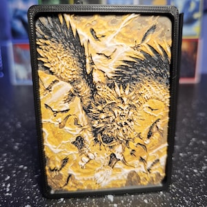 May include: A rectangular black plastic deck box featuring a textured, three-dimensional relief panel on the front. The panel displays a detailed golden-yellow and black illustration of a winged creature with sharp features and outstretched wings.