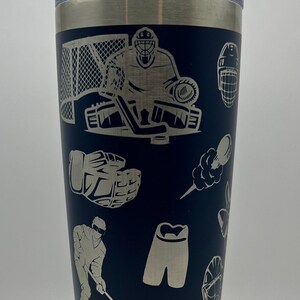 May include: A stainless steel tumbler with a navy blue exterior and a white design of hockey equipment, including a goalie, a stick, a puck, and a glove.