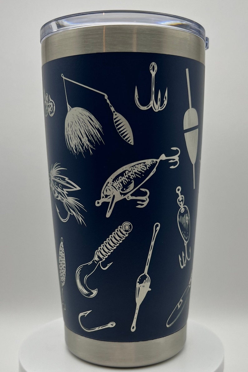 May include: Navy blue insulated tumbler with a silver rim and lid. The tumbler is decorated with a repeating pattern of fishing lures, hooks, and bobbers in a white, etched design. A great gift for anglers.
