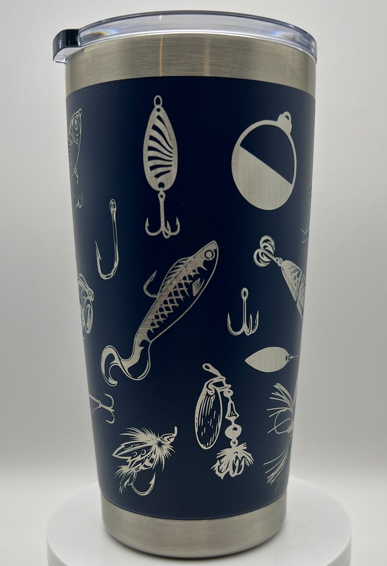 May include: Navy blue insulated tumbler with a stainless steel rim and lid. The tumbler is decorated with etched fishing lures, hooks, and a bobber. The design is repeated around the entire surface of the tumbler.