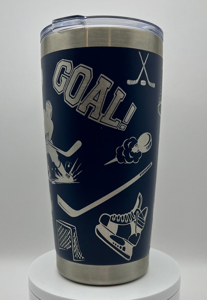 May include: A blue stainless steel tumbler with a silver lid. The tumbler is decorated with a hockey theme, including a hockey player, a hockey stick, a puck, and the word "GOAL!" in white lettering.