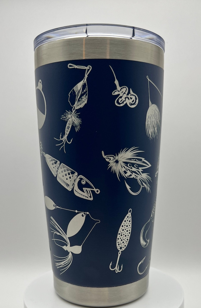 May include: Navy blue insulated tumbler with a silver rim and lid. The tumbler is decorated with a pattern of various fishing lures in a white, etched design. The tumbler is designed to keep beverages cold or hot.
