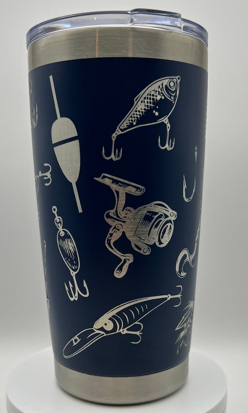 May include: Navy blue insulated tumbler with a silver lid and base. The tumbler is decorated with a pattern of fishing lures, hooks, and a fishing reel in a silver color. The tumbler is designed to keep beverages cold or hot.