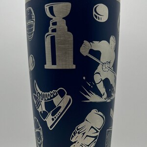 May include: A stainless steel tumbler with a navy blue exterior and a silver lid. The tumbler features a hockey-themed design with images of hockey players, pucks, skates, and a hockey trophy.