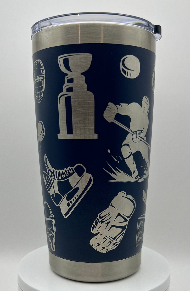 May include: A stainless steel tumbler with a navy blue exterior and a silver lid. The tumbler features a hockey-themed design with images of hockey players, pucks, skates, and a hockey trophy.