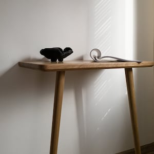 May include: A black ceramic bowl sits on a wooden table with a white book lying on the table. The table has two wooden legs and is in front of a white wall.