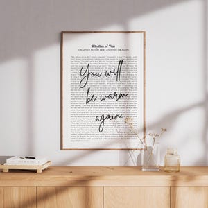 以下が含まれることがあります： 手書きの「You will be warm again」というテキストが書かれたフレーム付きプリント。プリントには「Rhythm of War, Chapter 80: The Dog and the Dragon」というテキストも含まれています。プリントは木製の表面に表示されています。