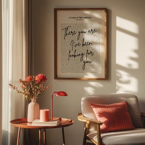 Op de afbeelding: Een ingelijste print met de tekst "There you are, I've been looking for you" uit "A Court of Mist and Fury". De print wordt weergegeven boven een bijzettafel met een rode lamp, een vaas met bloemen en een kaars. Naast de tafel staat een grijze fauteuil met een koraalkleurig kussen.