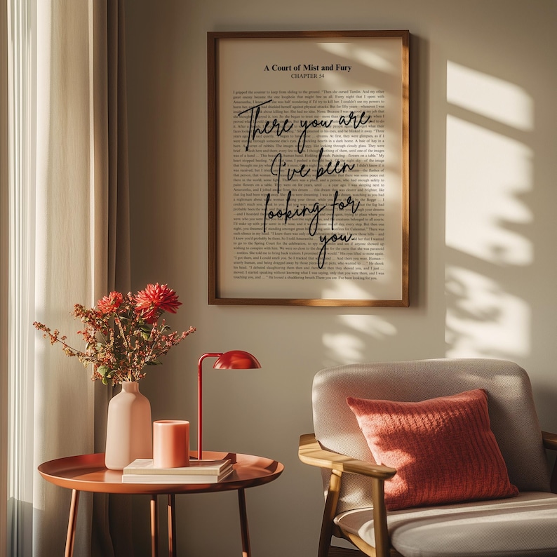 Op de afbeelding: Een ingelijste print met de tekst "There you are, I've been looking for you" uit "A Court of Mist and Fury". De print wordt weergegeven boven een bijzettafel met een rode lamp, een vaas met bloemen en een kaars. Naast de tafel staat een grijze fauteuil met een koraalkleurig kussen.