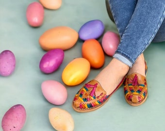 Pode incluir: Um par de sandálias de couro tecido colorido com borlas. As sandálias estão sobre um fundo azul claro com uma variedade de ovos de cores pastel.