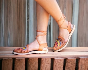 Puede incluir: Un par de sandalias de cuero marrón con bordados coloridos. Las sandalias tienen cordones que se atan al tobillo.