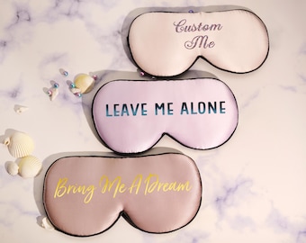 May include: Three sleep masks with black trim. The top mask is light pink with the text Custom Me. The middle mask is lavender with the text LEAVE ME ALONE in a blue gradient. The bottom mask is mauve with the text Bring Me A Dream in gold.