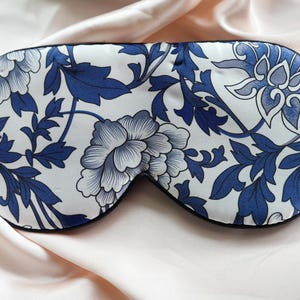 May include: A white sleep mask with a blue and white floral pattern. The mask is made of silk and has a black trim.