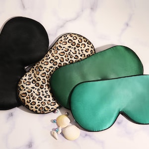 Peut inclure: Quatre masques de sommeil de différentes couleurs et motifs. Les masques sont faits d'un matériau doux et soyeux et ont une bordure noire. Les masques sont disposés en pile sur une surface en marbre blanc.