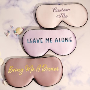 Könnte beinhalten: Drei Satin-Schlafmasken mit verschiedenen Sprüchen. Die obere Maske ist weiß mit schwarzem Besatz und sagt "Custom Me" in glitzernden pinkfarbenen Buchstaben. Die mittlere Maske ist hellrosa mit schwarzem Besatz und sagt "Leave Me Alone" in blauen Buchstaben. Die untere Maske ist hellbraun mit schwarzem Besatz und sagt "Bring Me A Dream" in goldenen Buchstaben.