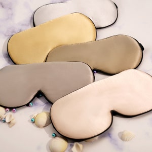 Pode incluir: Cinco máscaras de dormir de diferentes cores feitas de seda com acabamento preto. As máscaras estão empilhadas, com a cor mais clara na parte superior e a cor mais escura na parte inferior.