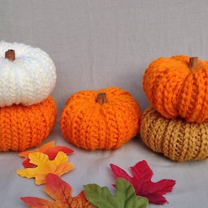 May include: Four crocheted pumpkins in orange, white, and brown. The pumpkins are stacked on top of each other. There are fall leaves scattered around the pumpkins.