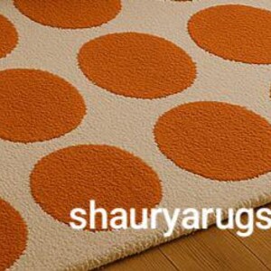 Peut inclure: Un tapis cr&egrave;me avec de grands pois orange. Le tapis a une surface textur&eacute;e et est pr&eacute;sent&eacute; sur un sol en bois. Le texte "shauryarugs" est visible sur le tapis.