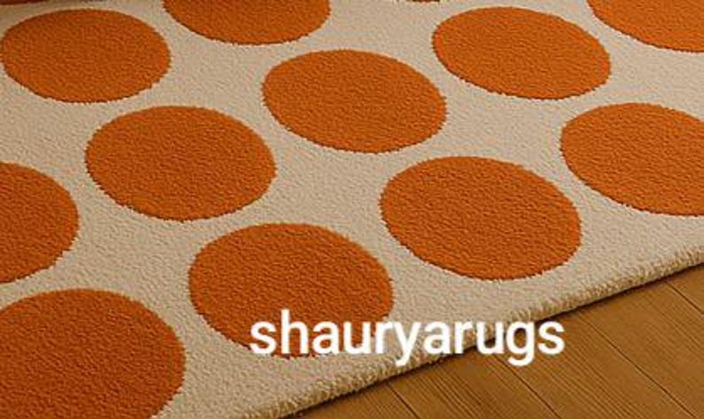 Peut inclure: Un tapis cr&egrave;me avec de grands pois orange. Le tapis a une surface textur&eacute;e et est pr&eacute;sent&eacute; sur un sol en bois. Le texte "shauryarugs" est visible sur le tapis.