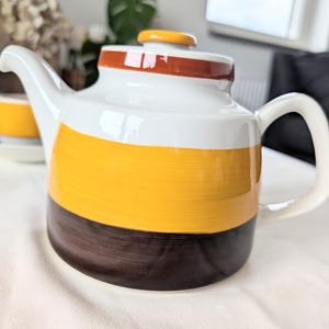 Op de afbeelding: Een keramische theepot met horizontale banden in wit, felgeel en donkerbruin. De theepot heeft een gebogen handvat, een tuit en een bijpassend deksel met een gele knop. Het oppervlak vertoont zichtbare texturen van penseelstreken in de verf.
