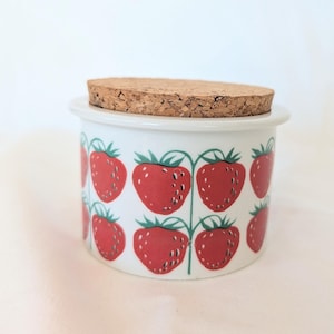 May include: A cylindrical white ceramic jar featuring a repeating pattern of red strawberries with green stems and leaves. The container is topped with a circular, textured brown cork lid that fits securely into the rim of the ceramic vessel.