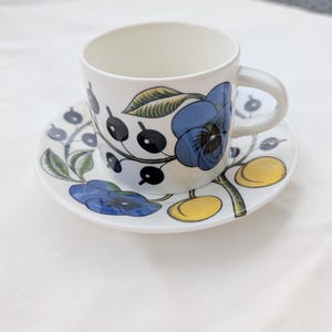 Könnte beinhalten: Ein weißes Tassen- und Untertassen-Set aus Keramik mit einem botanischen Muster aus blauen Stiefmütterchen, gelben Früchten und schwarzen Beeren an grünen Stielen. Die Tasse hat einen seitlichen Henkel und eine glatte Oberfläche mit floralem Dekor.