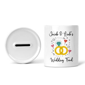 May include: White ceramic piggy bank with a slot on top. The front of the bank has a design of two gold rings with a diamond, red hearts, and the text "Jacob & Leah's Wedding Fund".