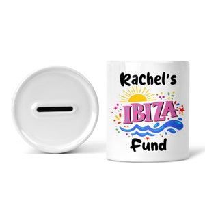 May include: White ceramic piggy bank with a slot on the top. The front of the bank has a colorful design with a sun, waves, and the text "Rachel's Ibiza Fund".