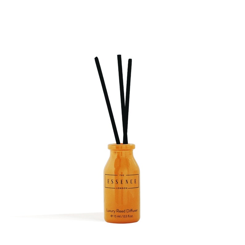May include: An orange ceramic reed diffuser with three black reeds. The bottle has a label that reads "THE ESSENCE LONDON" and "Luxury Reed Diffuser e 15 ml / 0.5 fl.oz.". The diffuser is set against a white background.