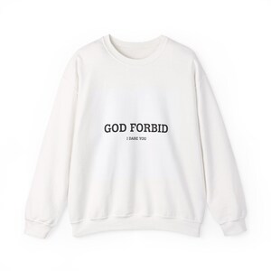 May include: A white crew neck sweatshirt with the text "GOD FORBID" above "I DARE YOU" in black. The sweatshirt has long sleeves and a relaxed fit, suitable for casual wear.