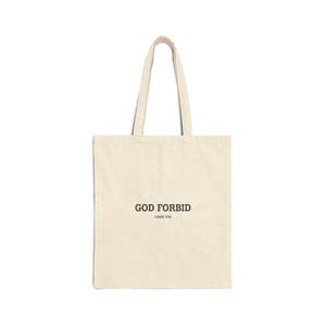 May include: Cream-colored tote bag with black text that reads "GOD FORBID" above "I DARE YOU". The bag has two handles and is made of a sturdy fabric, suitable for carrying books, groceries, or other items.